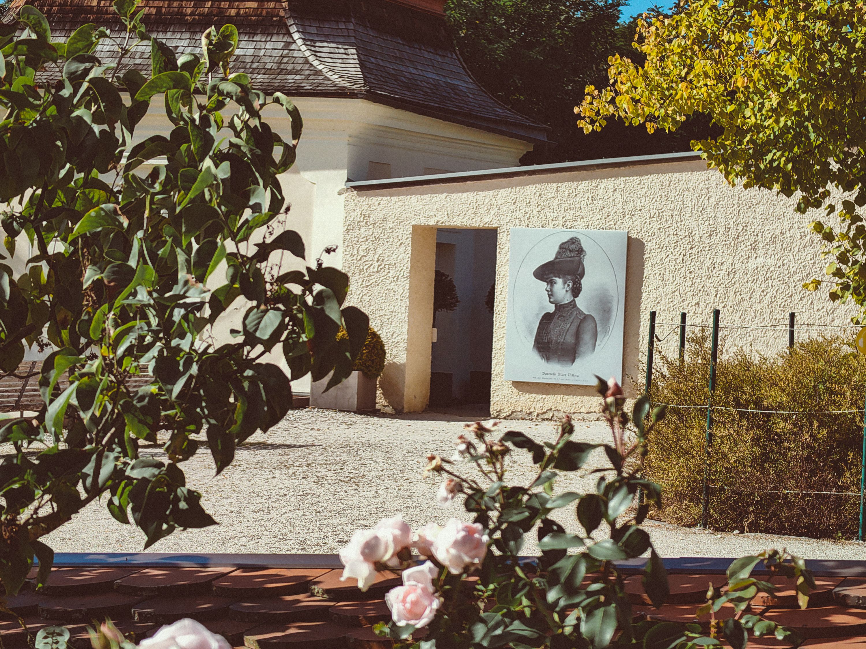 Umgeben von üppigem Grün und blühenden Rosen entfaltet sich hier eine Oase der Ruhe. Der eindrucksvolle Wandgemälde an der Wand erzählt Geschichten aus der Vergangenheit und lädt Besucher ein, die spirituelle Atmosphäre dieses besonderen Ortes zu erleben.