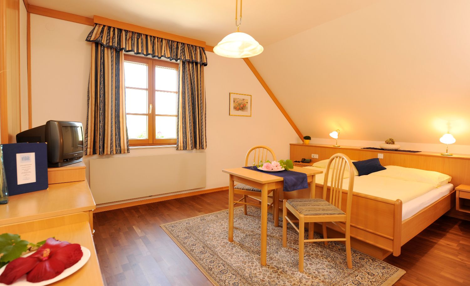 Hangulatos vendégszoba fabútorokkal, franciaággyal, kis asztallal és székekkel, TV-vel és függönyös ablakkal.