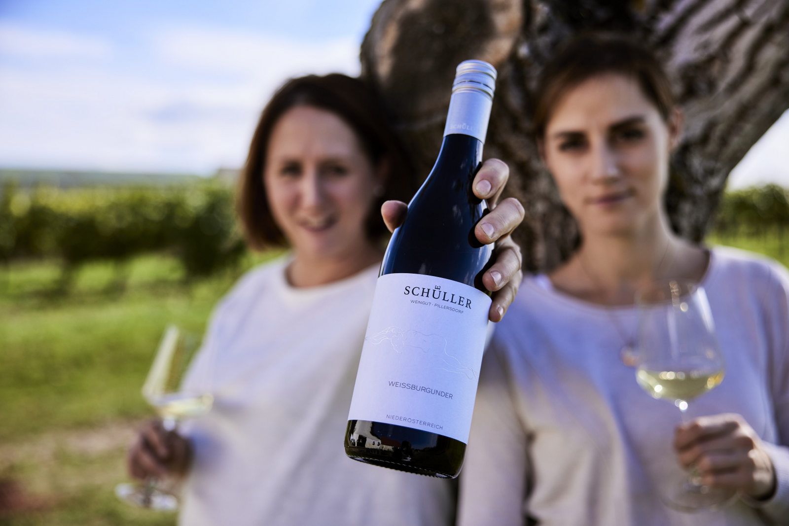 Két ember egy üveg Schüller Pinot Blanc-t és borospoharakat tart egy szőlőskertben egy fa előtt.