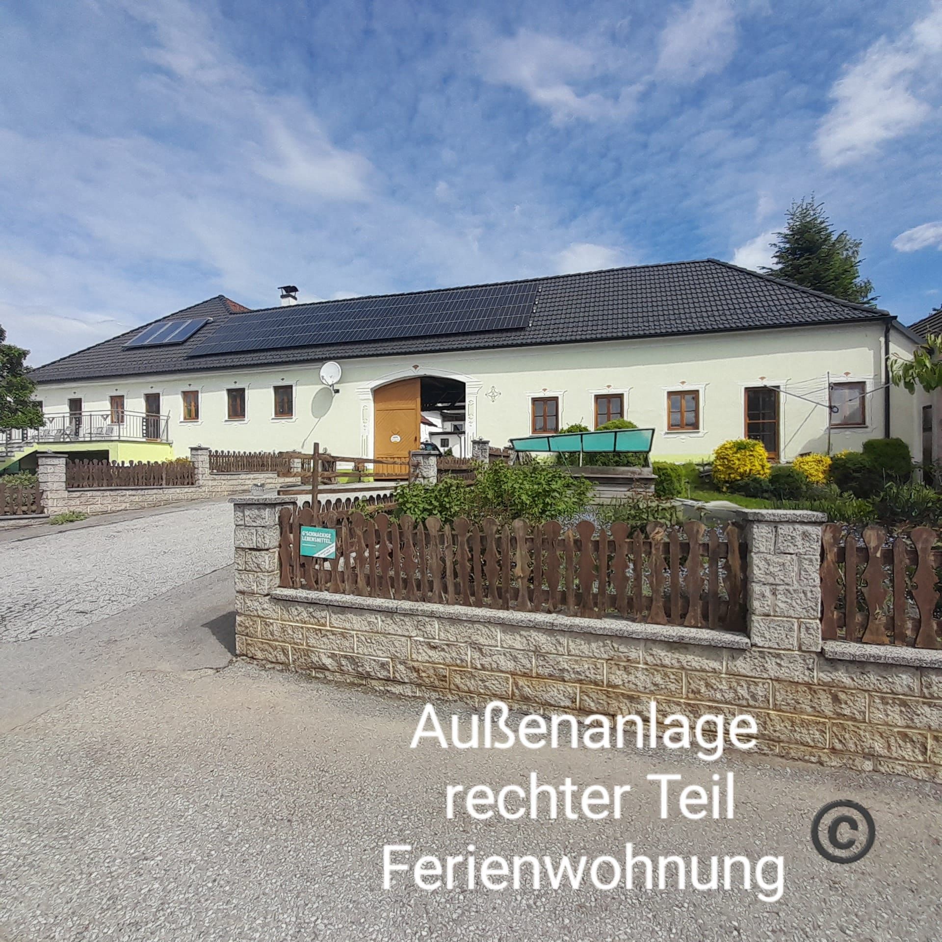 Egy nagy épület sárga homlokzattal és fekete tetővel, melyet kert és fakerítés vesz körül. Az ég kék, néhány felhővel.