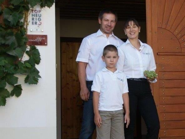 Egy család áll egy faajtós épület előtt, a nő szőlővel a kezében.