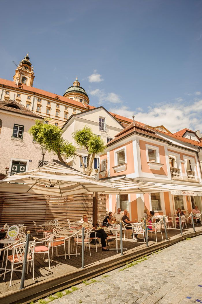 Egy kávézó szabadtéri területe napernyőkkel és vendégekkel, a háttérben egy történelmi épület kupolával.