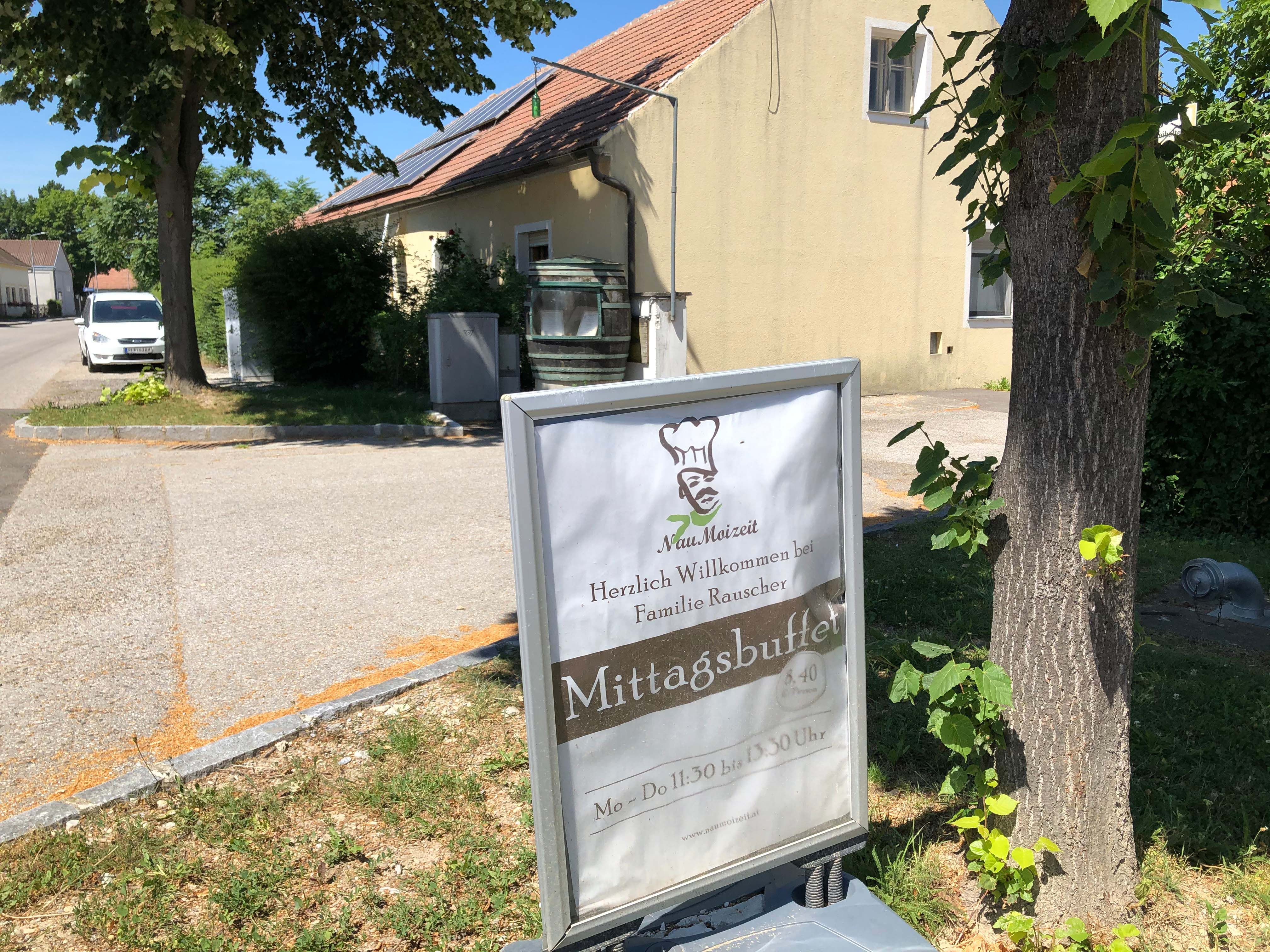 Egy épület előtti tábla a "Mittagsbuffet" és a "Herzlich Willkommen bei Familie Rauscher" felirattal.
