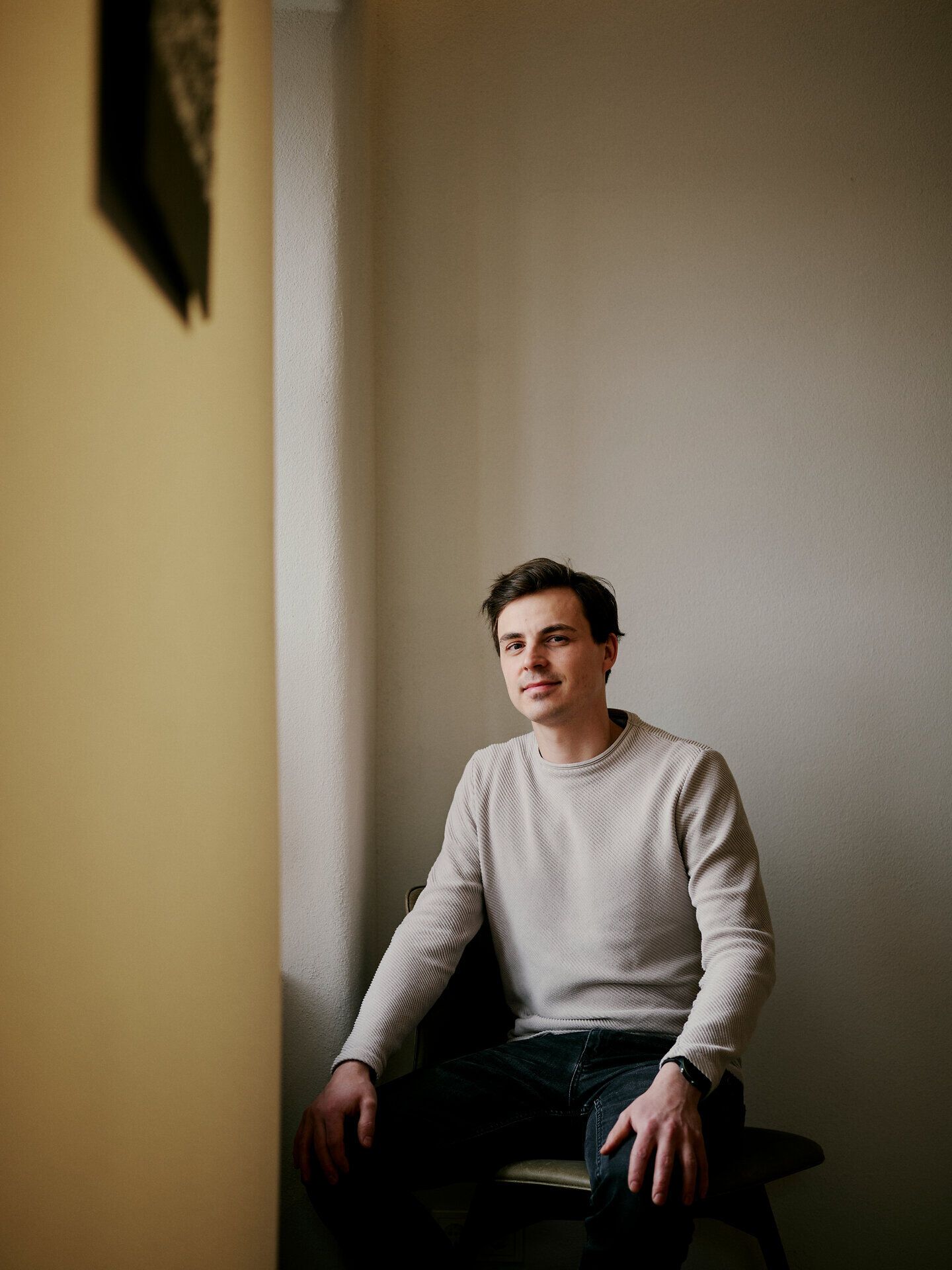 Egy személy egy sámlin ül egy egyszerű belső térben, és a kamerába néz.