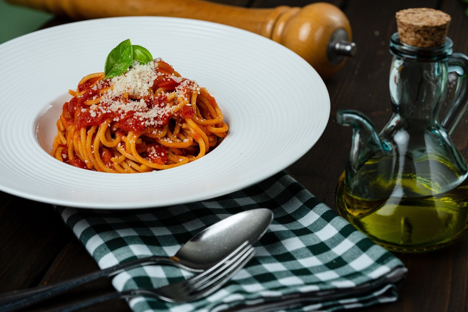 Egy tányér spagetti paradicsomszósszal, bazsalikommal és parmezánnal, mellette egy üveg olaj és evőeszközök egy kockás ruhán.
