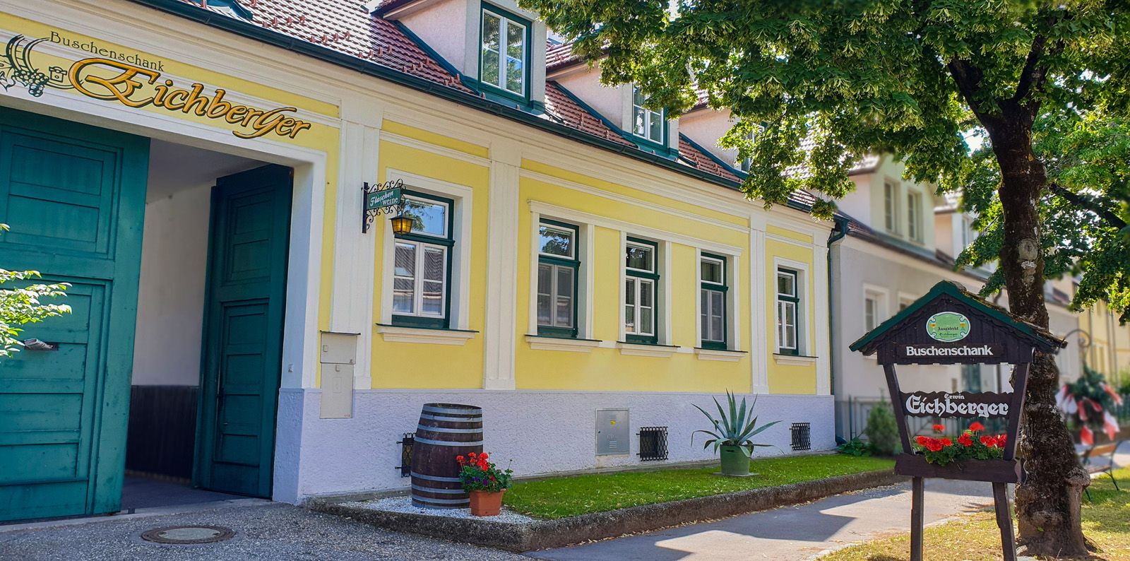 Sárga épület zöld ajtókkal és ablakokkal, "Hagyományos borozó Eichberger" felirat, előtérben fa.