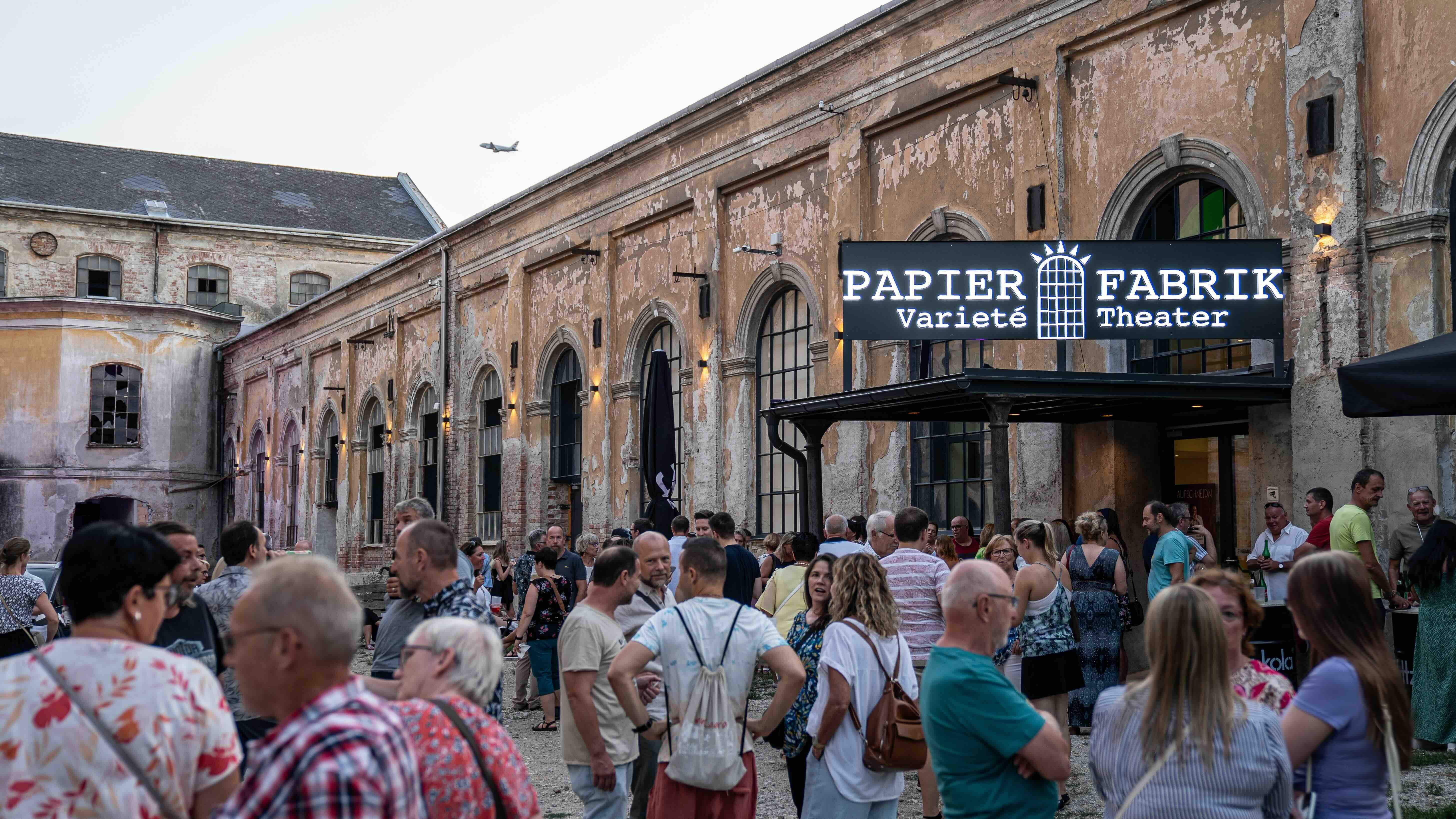 Tömeg egy régi papírgyár előtt a "Papier Fabrik Varieté Színház" felirattal.