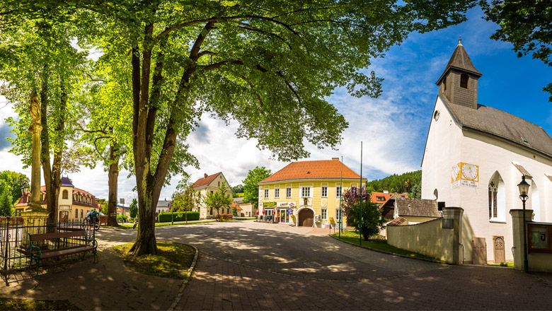 Panorámakép a Feistritz templomtérről a templommal, fákkal és történelmi épületekkel.