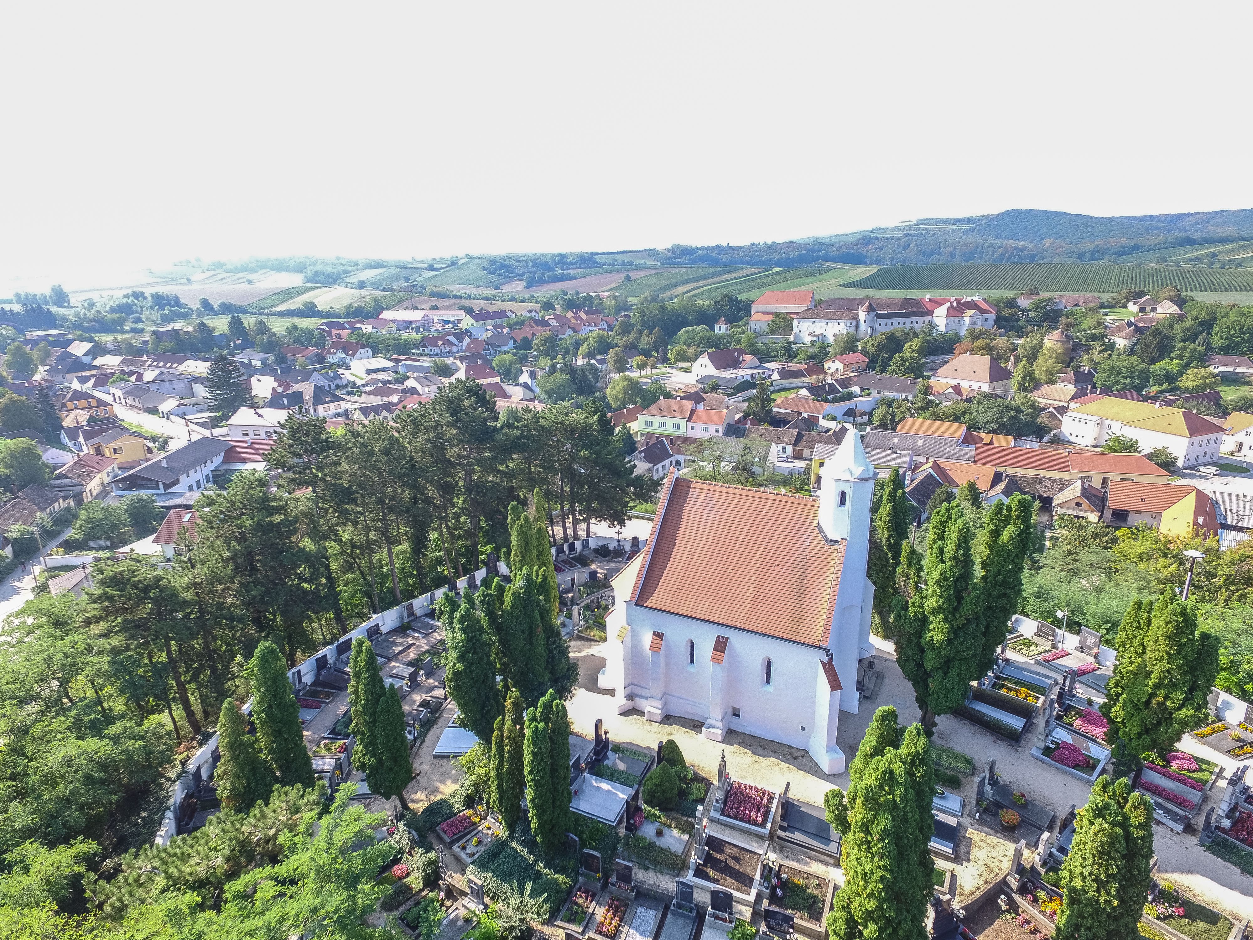 Légi felvétel egy piros tetejű templomról, amelyet temető és fák vesznek körül, vidéki környezetben, a háttérben házakkal és dombokkal.