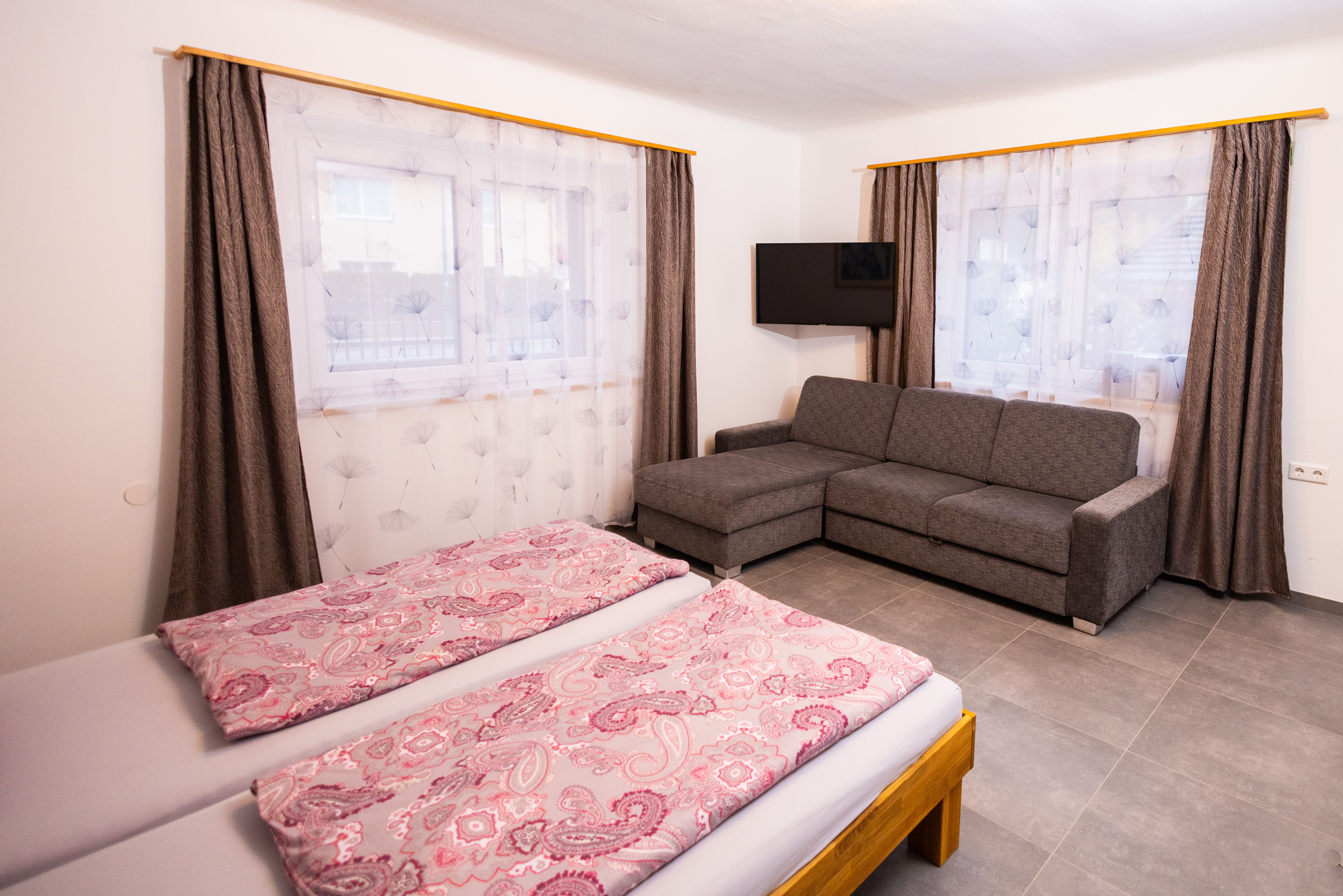 Egy hálószoba franciaággyal, szürke kanapéval, TV-vel és két függönyös ablakkal.