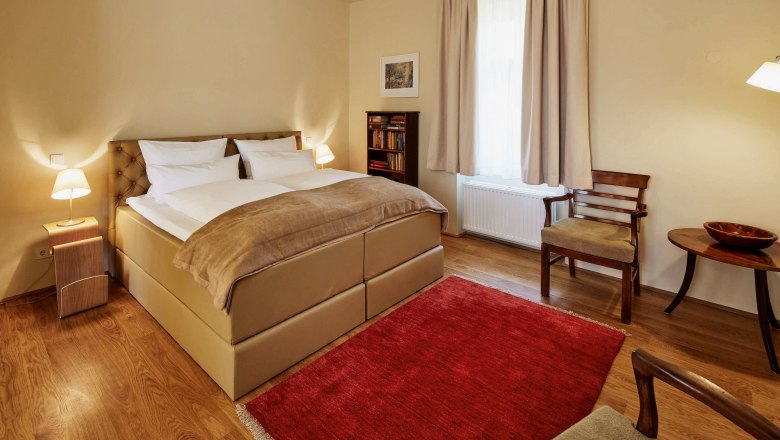 Hangulatos hálószoba nagy ággyal, két éjjeliszekrénnyel, lámpákkal, piros szőnyeggel és fabútorokkal.