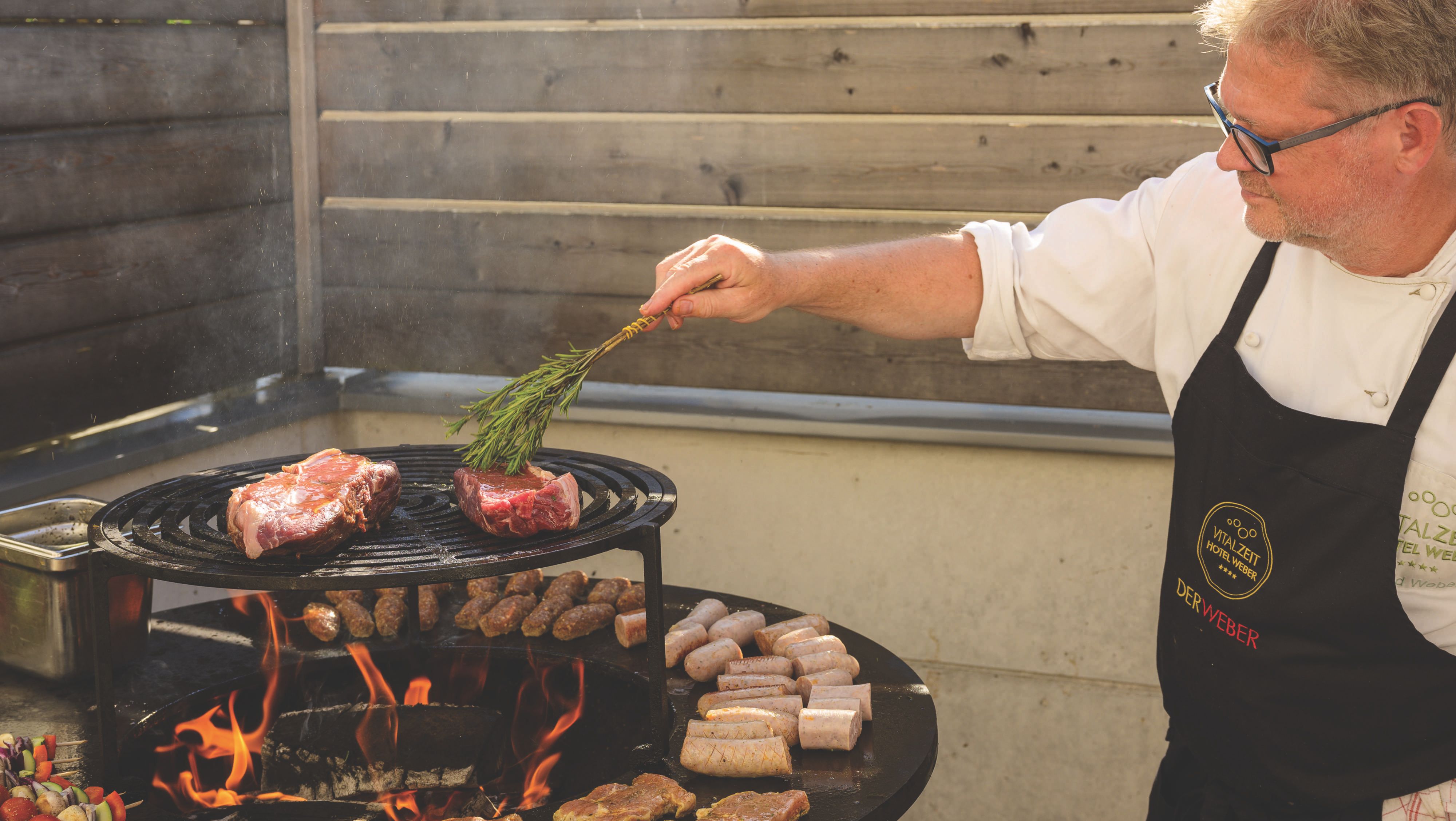 A ház ura nyílt tűzön, egy kerek grillen süt húst és kolbászt.